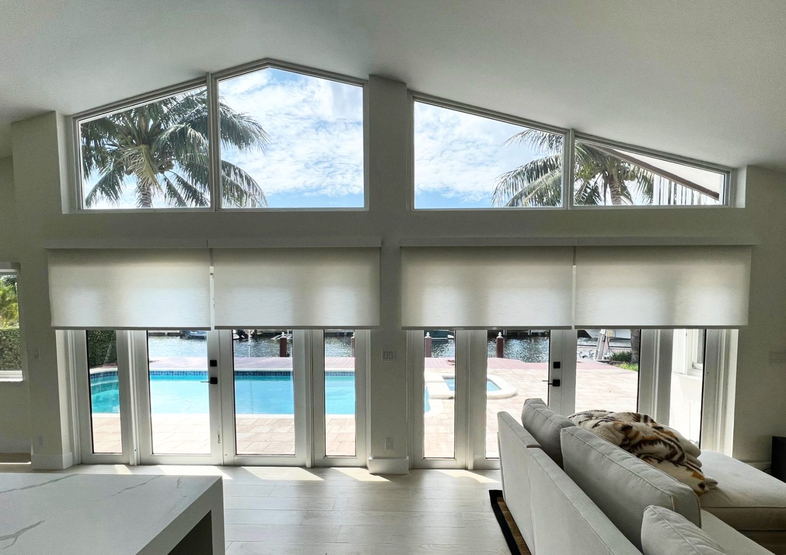 Ivory roller shades across a vaulted waterfront wall, sized so the gable glass above stays open to the sky.