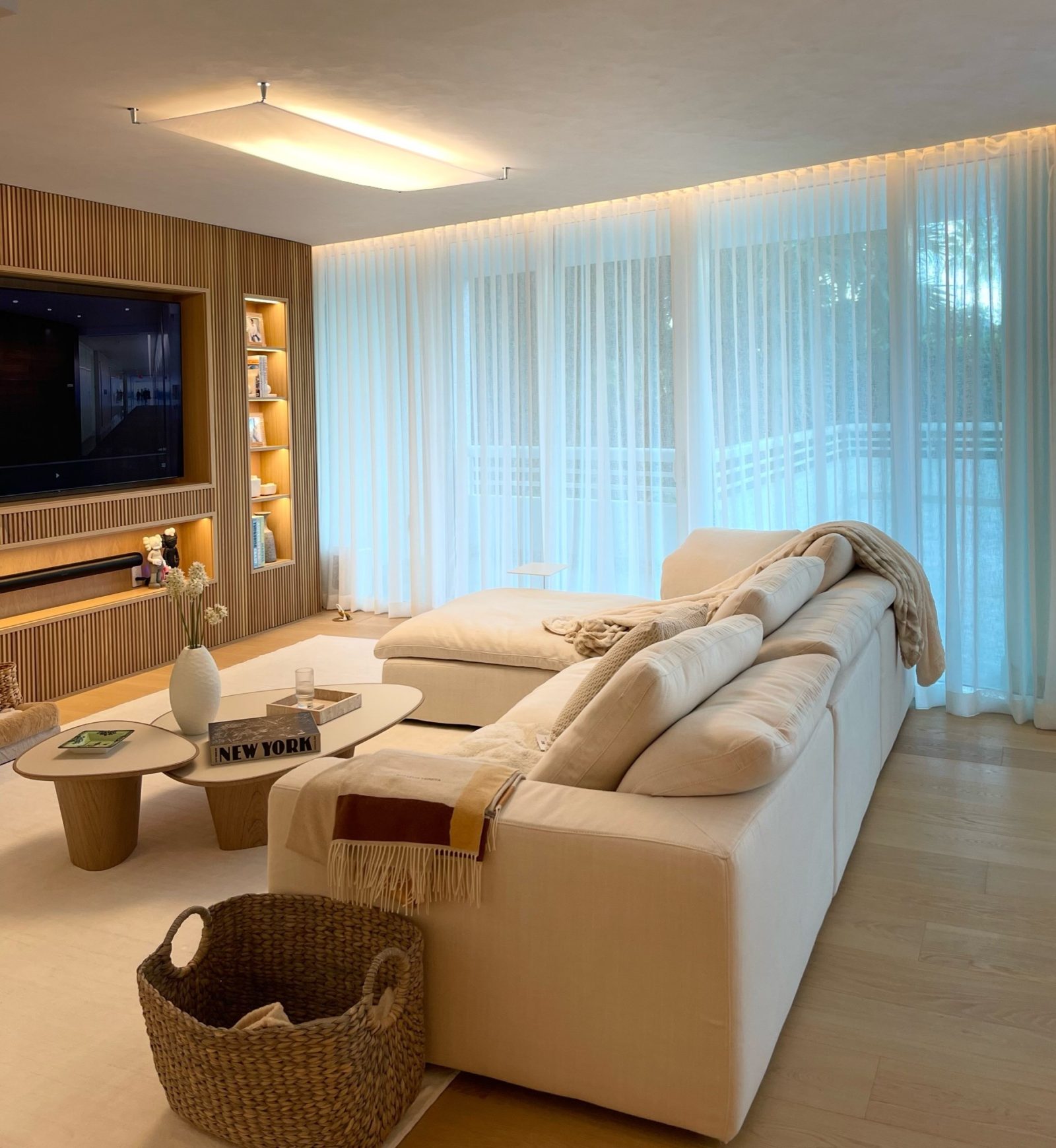 A backlit wall of sheer drapery beside a slatted wood media wall, lit from a recessed cove above the track.
