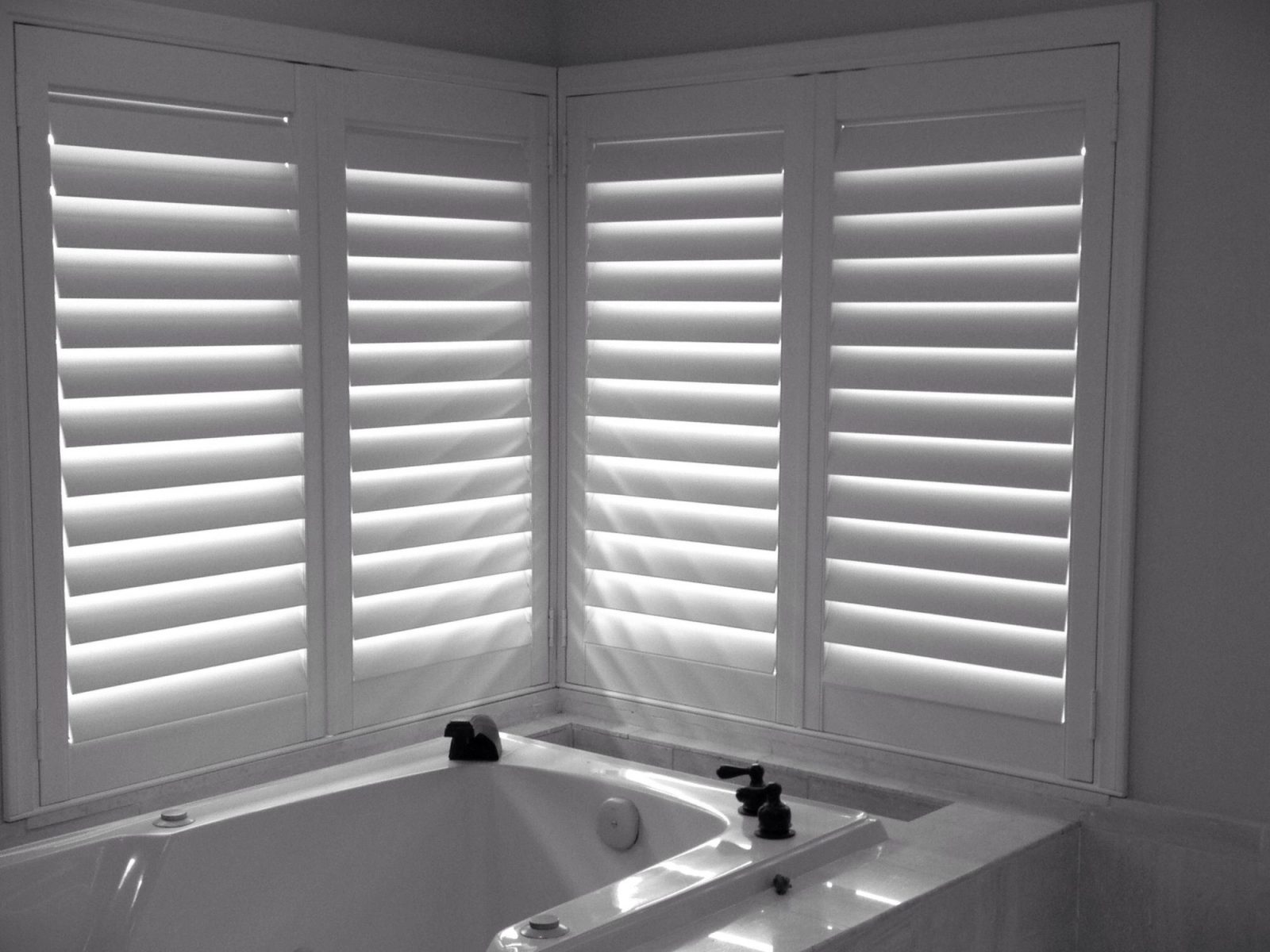 White shutters wrapped into a bathroom corner above a tub, louvers angled up for privacy and daylight.
