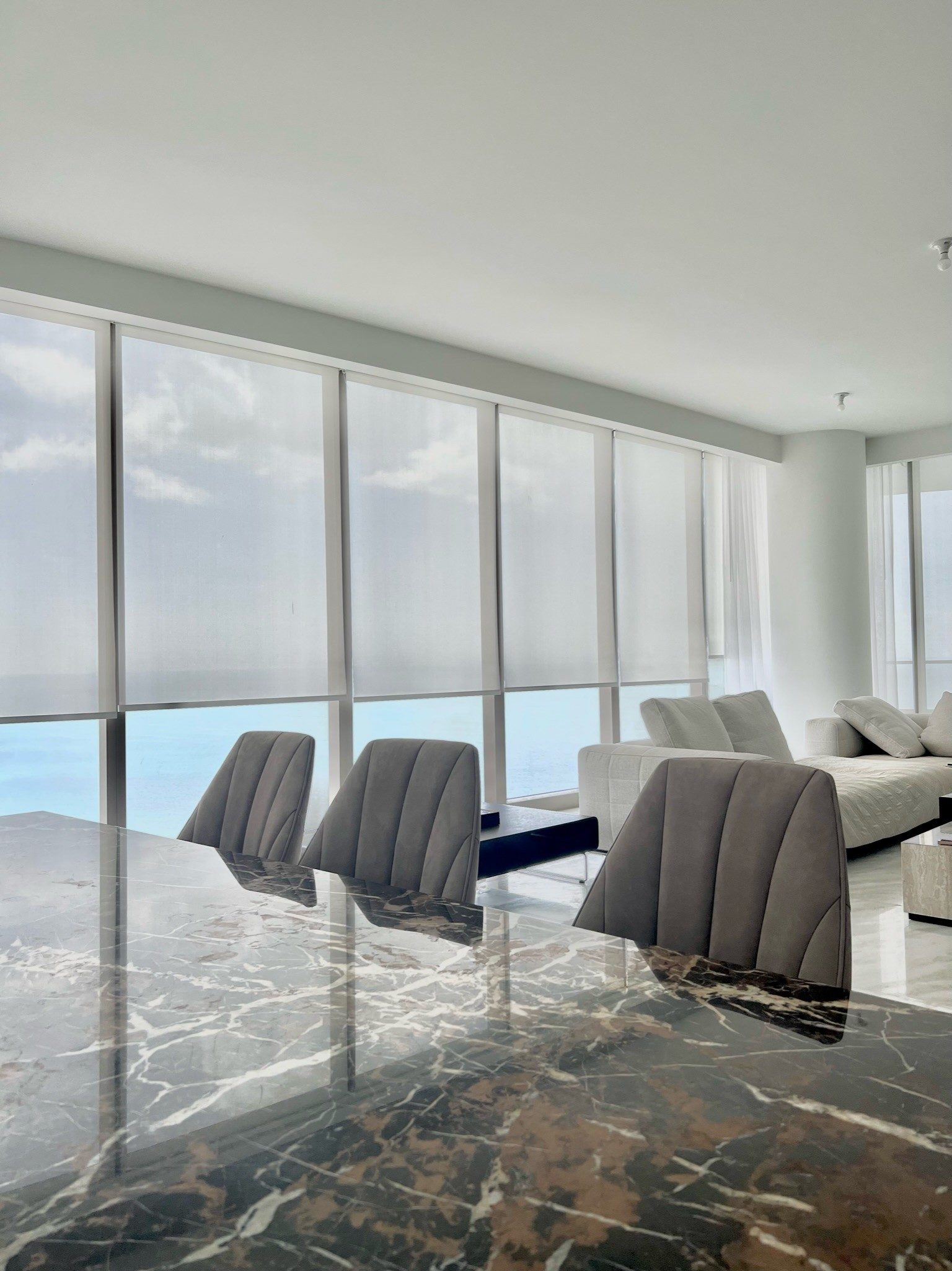Solar shades on an oceanfront wall, dropped to the sill line so the horizon stays in view from the table.